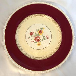 Pode incluir: Um prato decorativo com uma borda bordô e um centro creme. O centro apresenta um desenho floral com rosas vermelhas e amarelas. O prato tem uma borda branca ondulada e acabamentos dourados, mostrando uma estética vintage.
