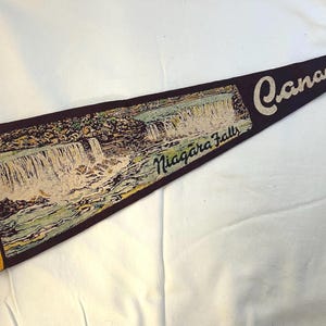 May include: A brown felt pennant with a yellow border and a picture of Niagara Falls. The pennant has the word "Canada" written in white on a dark brown background.