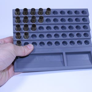 May include: A grey 9mm ammunition loading tray with multiple circular holes for brass casings. The tray is rectangular with two rectangular compartments at the bottom. The tray is held by a hand.