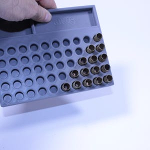 May include: A gray plastic ammunition loading tray with multiple circular holes, some filled with brass casings. The tray is held by a hand and is labeled "9mm". The background is white.
