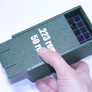 May include: Green rectangular storage box with a sliding lid, labelled "223 rem 50 rds." The box is open, revealing a grid of compartments, one of which holds a bullet. The box is held by a hand.