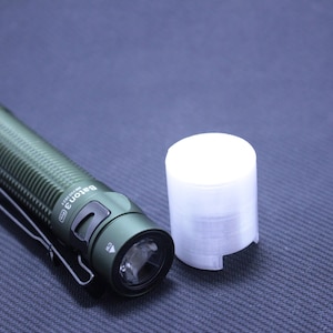 Könnte beinhalten: Eine olivgrüne Taschenlampe mit Taschenclip und weißer zylindrischer Abdeckung. Auf der Taschenlampe steht "Baton 3". Die Taschenlampe liegt auf einer dunkelblauen Oberfläche.