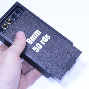 Può includere: Una scatola nera per la conservazione di munizioni da 9 mm con la scritta bianca "9mm 50 rds". La scatola, tenuta in mano, contiene due proiettili color oro. La scatola ha una superficie strutturata.