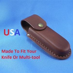 May include: Brown leather knife or multi-tool sheath with a snap closure. The sheath is stitched around the edges and has a loop for a belt. The text "USA" is in red and blue. The text "Made To Fit Your Knife Or Multi-tool" is in red.