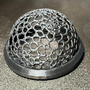May include: A black, 3D-printed dome-shaped object with a honeycomb-like pattern. The object sits on a circular base. The intricate design and dark color give it a modern, industrial aesthetic. The object is likely a decorative or functional item.