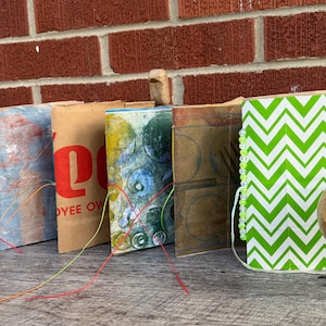May include: A collection of handmade journals and decorative wooden bird sculptures. The journals feature various covers, including patterned paper and repurposed materials. The birds have wire legs and colorful wire accents, adding a whimsical touch to the display.