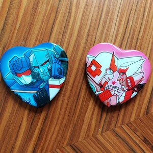 Könnte beinhalten: Zwei herzförmige Buttons mit Cartoon-Charakteren. Einer ist blau mit zwei Charakteren, der andere rosa mit zwei Charakteren. Die Buttons liegen auf einer Holzoberfläche.