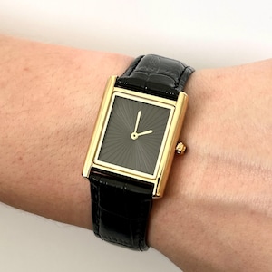 Peut inclure: Une montre-bracelet rectangulaire dorée avec un cadran et des aiguilles noirs, et un bracelet en cuir noir. Le cadran de la montre présente un motif en forme d'éclat de soleil. La montre mesure environ 3,8 cm de long.