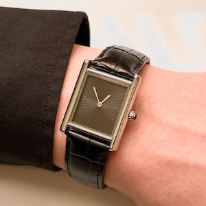 Könnte beinhalten: Eine rechteckige Armbanduhr mit schwarzem Zifferblatt und silberfarbenem Gehäuse. Die Uhr hat ein schwarzes Lederarmband. Die Uhr befindet sich am Handgelenk einer Person, neben einem schwarzen Ärmel.