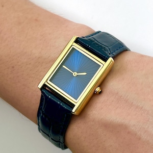 Peut inclure: Une montre-bracelet rectangulaire dorée avec un cadran bleu en forme de soleil et des aiguilles dorées. La montre a un bracelet en cuir bleu foncé. La montre est sur un poignet.