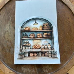 May include: Watercolor illustration of a bakery scene with various types of bread on shelves. The image features a light blue archway, a hanging light, and baskets of baked goods. The artwork is set on a wooden tray.