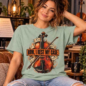 Könnte beinhalten: Ein salbeigrünes T-Shirt mit einer Grafik einer Geige und eines Bogens vor einem Hintergrund aus Noten. Ein rechteckiges orangefarbenes Banner in der Mitte enthält den schwarzen Text "BOW, I REST MY CASE" in einer fetten Schriftart.