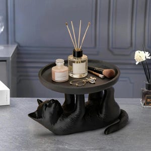 May include: A black cat-shaped decorative tray, holding a small bottle, a diffuser, makeup brushes, and jewelry. The tray is round and dark gray. The cat is lying on its back, supporting the tray with its paws.