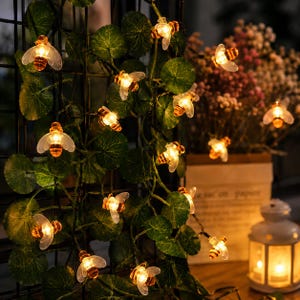 May include: A string of decorative LED lights shaped like small bees with translucent wings and glowing amber bodies, intertwined with artificial green leaves on a dark wire frame, creating a warm illumination effect for indoor decor.