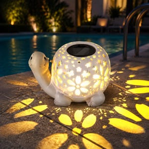 May include: A white turtle-shaped solar garden light with a shell featuring intricate cut-out flower and star patterns. The light emits a warm yellow glow, casting petal-shaped shadows on the ground. A circular solar panel is visible on top.