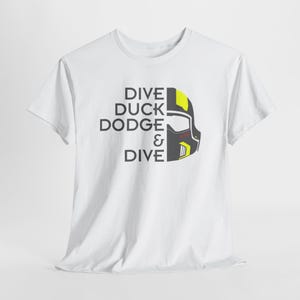 May include: White t-shirt with the words "DIVE DUCK DODGE & DIVE" in black text. A graphic of a helmet with yellow and red accents is on the right side of the text. The t-shirt is made of a soft material.