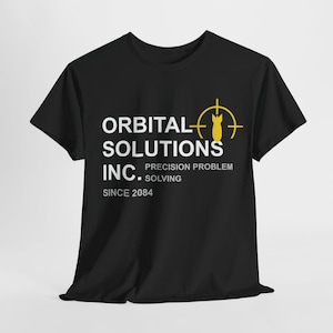 May include: Black t-shirt featuring the white text "ORBITAL SOLUTIONS INC." and a yellow bomb graphic within a target. The shirt also displays "PRECISION PROBLEM SOLVING" and "SINCE 2084".