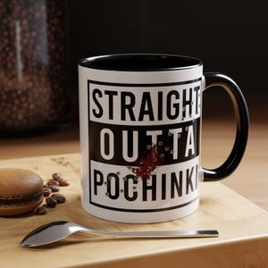 May include: White ceramic mug with a black rim and handle. The mug features a black and white graphic that says "STRAIGHT OUTTA POCHINKI" with a red splatter and bullet holes.