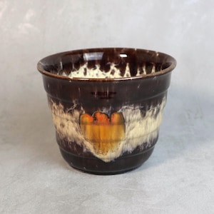Könnte beinhalten: Ein brauner Keramik-Blumentopf mit einem einzigartigen Design. Der Topf hat eine dunkelbraune Außenseite, ein cremefarbenes Band und ein zentrales Design mit orangefarbenen und gelben Farbtönen. Der Rand ist glänzend. Durchmesser ca. 15 cm, für drinnen und draußen geeignet.