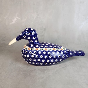 Könnte beinhalten: Keramik-Entenfigur mit blau-weißem Tupfenmuster. Die Ente hat einen weißen Schnabel und ein dekoratives Band mit orangefarbenen, grünen und weißen Blumenmustern. Ein dekorativer Artikel für das Zuhause.