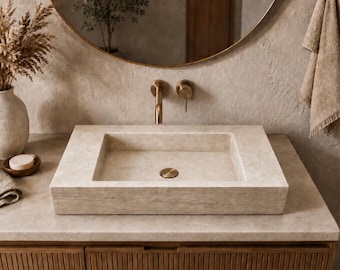 May include: A rectangular, light-colored stone sink with a brass drain and faucet. The sink is set on a wooden vanity with a matching countertop. A round mirror and a vase with dried flowers are visible in the background.