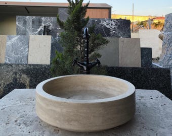 May include: A round, beige stone sink with a black faucet. The sink is set on a gray stone surface. Behind the sink is a small evergreen tree and a display of various stone tiles.