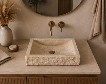 May include: A beige stone bathroom sink with a square basin and rough-hewn edges. The sink is set on a light-colored countertop with a wooden cabinet below. A gold faucet and mirror are mounted on the wall, with a vase of dried flowers and a towel nearby.