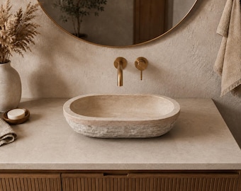 May include: A bathroom vanity with a stone-like oval sink, gold fixtures, and a large round mirror. A vase with dried plants and a small soap dish are on the countertop. The vanity has a wooden cabinet.