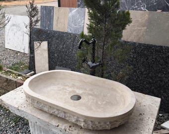 May include: A beige, oval-shaped stone sink with a dark faucet. The sink is set on a matching stone base. Various stone slabs are in the background, with a small tree to the right of the faucet. The sink has a drain hole.