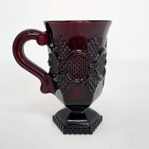 以下が含まれることがあります： 装飾模様のルビーレッドガラスマグ。マグにはハンドルと台座が付いています。デザインには、ダイヤモンドと円形のパターンが含まれています。マグの高さは約13cmです。