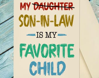 May include: A greeting card with the text "My Son-In-Law Is My Favorite Child" in red, gold, green, and blue. The word "Daughter" is crossed out. The card is cream-colored and comes with a matching envelope.