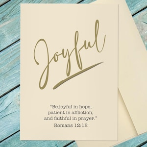 May include: A cream-colored greeting card featuring the word Joyful in large gold cursive script with a gold underline. Below the text is the quote Be joyful in hope, patient in affliction, and faithful in prayer. Romans 12:12 in black font.