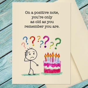 May include: A birthday greeting card with a humorous illustration. The card features a stick figure looking confused next to a cake with lit candles. The text reads, "On a positive note, you're only as old as you remember you are."
