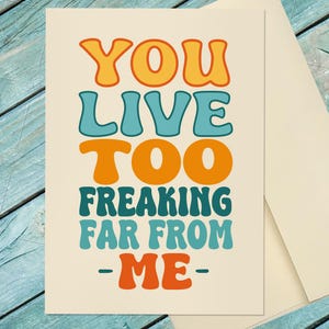 May include: A cream-colored greeting card with the text "YOU LIVE TOO FREAKING FAR FROM - ME -" in orange and teal retro font. The card is paired with a cream-colored envelope.