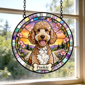 May include: Round stained glass window art featuring a poodle with a happy expression, framed by pink flowers and a colorful border. The dog's name, "Pookie," is on a bone-shaped tag. The artwork hangs from a black chain.