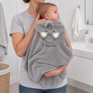 Puede incluir: Una toalla de bebé con capucha de color gris suave que presenta un diseño de elefante en la capucha con ojos y orejas bordados. La toalla está personalizada con el nombre Charlie bordado en hilo gris oscuro en el centro de la parte frontal.