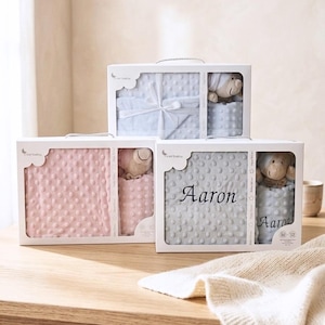 Peut inclure: Trois boîtes cadeaux rectangulaires blanches contenant des ensembles pour bébé, chacun comprenant une couverture minky à pois et une petite peluche. Une boîte affiche le prénom Aaron brodé en noir sur une couverture et un accessoire assorti.