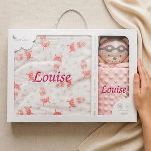 May include: A baby gift set in a white box, featuring a pink and white blanket with a repeating pattern of umbrellas and bears, and a matching pink bear plush toy. The name "Louise" is embroidered on both items.