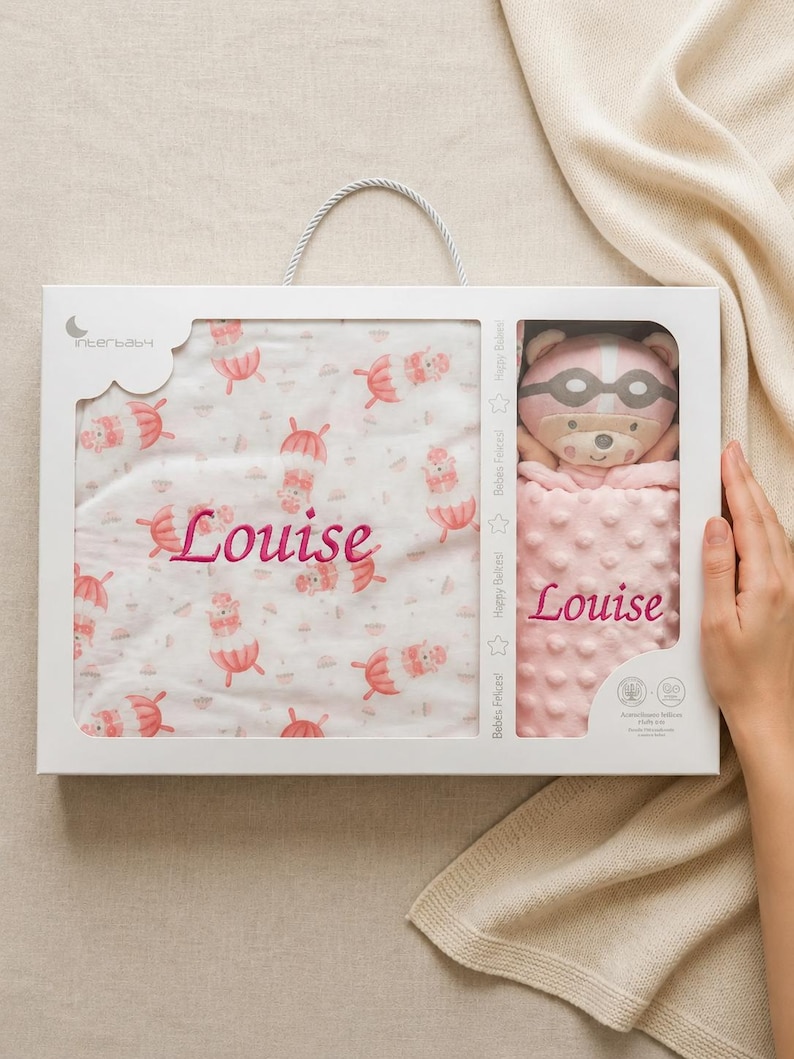 May include: A baby gift set in a white box, featuring a pink and white blanket with a repeating pattern of umbrellas and bears, and a matching pink bear plush toy. The name "Louise" is embroidered on both items.