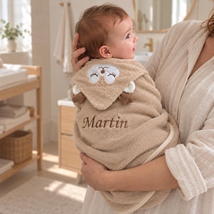 Peut inclure: Une serviette de bain à capuche pour bébé de couleur beige, ornée d'un visage d'animal brodé sur la capuche avec de petites oreilles et le prénom Martin cousu en fil marron sur le devant. La serviette est en tissu doux avec un liseré clair.