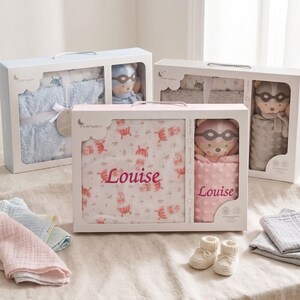May include: Gift sets for babies, each in a white box with a clear window. Sets include a blanket with the name "Louise", a plush toy, and other baby items. The blankets have a pattern of umbrellas and the toys are wearing masks.