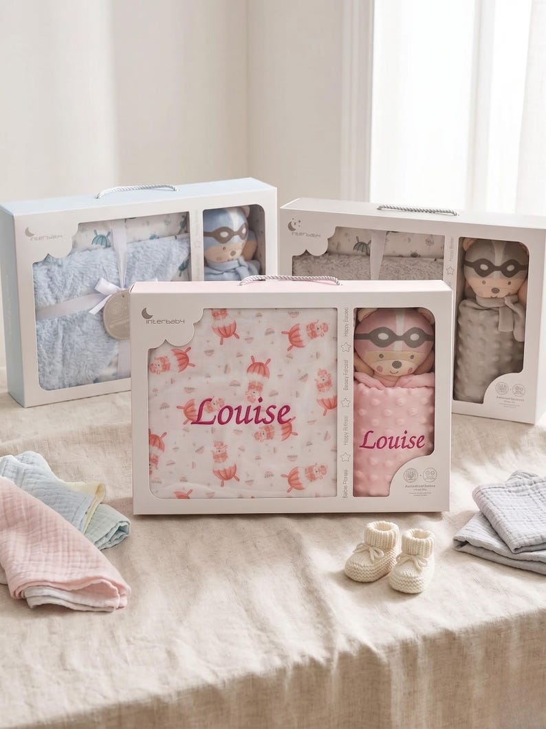 May include: Gift sets for babies, each in a white box with a clear window. Sets include a blanket with the name "Louise", a plush toy, and other baby items. The blankets have a pattern of umbrellas and the toys are wearing masks.