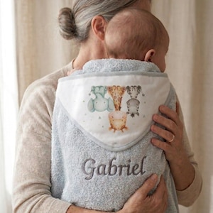 May include: A light blue hooded baby towel featuring a white hood lining with a printed illustration of an elephant, a giraffe, and a zebra. The name Gabriel is embroidered in dark gray thread on the main body of the soft, textured towel.