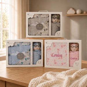 May include: Gift sets for babies, each in a white box with a clear window. The sets include a blanket, a soft toy, and other baby items. The blankets are in various colors, including gray, blue, and pink, with the name "Louise" on one.