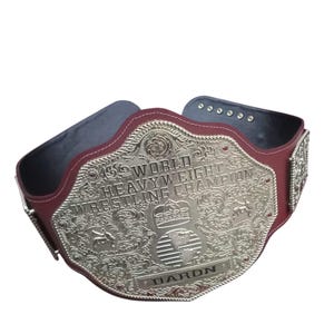 Puede incluir: Un cinturón de campeonato de lucha libre con una gran placa central de metal plateado ornamentada con volutas intrincadas, una corona, un globo terráqueo y el texto "WORLD HEAVYWEIGHT WRESTLING CHAMPION" y "DARON" sobre una correa de cuero granate.