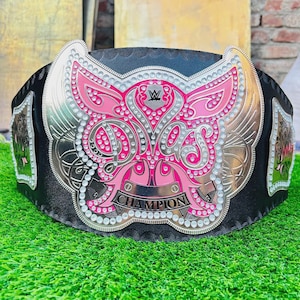 Könnte beinhalten: Ein schwarzer Leder-Meisterschaftsgürtel mit einer großen, verzierten silberfarbenen Metallplatte, die mit rosa Emaille-Akzenten und klaren Strasssteinen besetzt ist. Die Platte zeigt das Wort DIVAS und das Wort CHAMPION auf einem Banner.