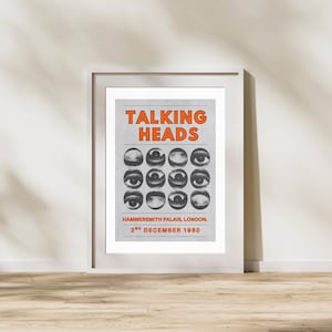 Op de afbeelding: Ingelijste poster van de band Talking Heads. De naam van de band staat in oranje letters bovenaan. De achtergrond is lichtgrijs met twaalf oogillustraties. Locatie en datum staan onderaan afgedrukt. De lijst is wit.