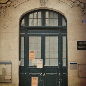 May include: An aged stone building with a dark green wooden door. The arched doorway has a transom window above the door. The words "ECOLE DE GARCONS" are carved above the door. Several posters are affixed to the door.