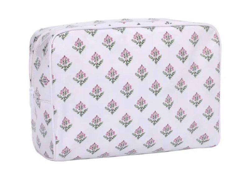 Op de afbeelding: Een witte make-uptas met een herhaald patroon van roze en groene bloemmotieven. De rechthoekige tas heeft afgeronde hoeken en een ritssluiting. Het bloemmotief is gelijkmatig verdeeld over het oppervlak.