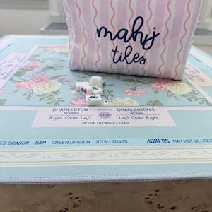 Op de afbeelding: Een roze en witte gestreepte mahjongtegeltas met de woorden "mahj tiles" in marineblauw. De tas staat op een mahjongtafel met bloemenpatroon en tegels. De tafel heeft de tekst "Charleston 1" en "Charleston 2".